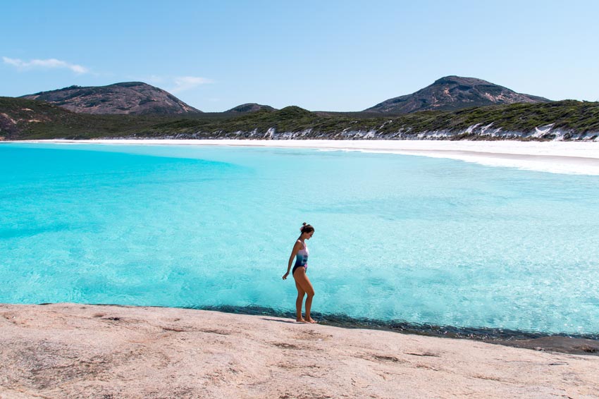una chica caminando en una gran roca frente a agua increíblemente azul y un largo arco de arena