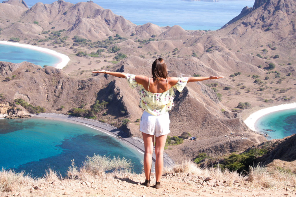una chica de pie en la parte superior de la isla de Padar con los brazos extendidos viendo hacia abajo en tres playas