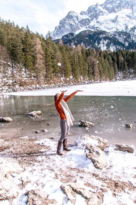 una chica con un suéter naranja arroja nieve en el aire con un lago y árboles en el fondo