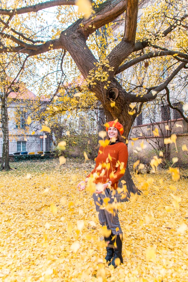 mujer con suéter naranja y sombrero lanzando hojas naranjas en Praga