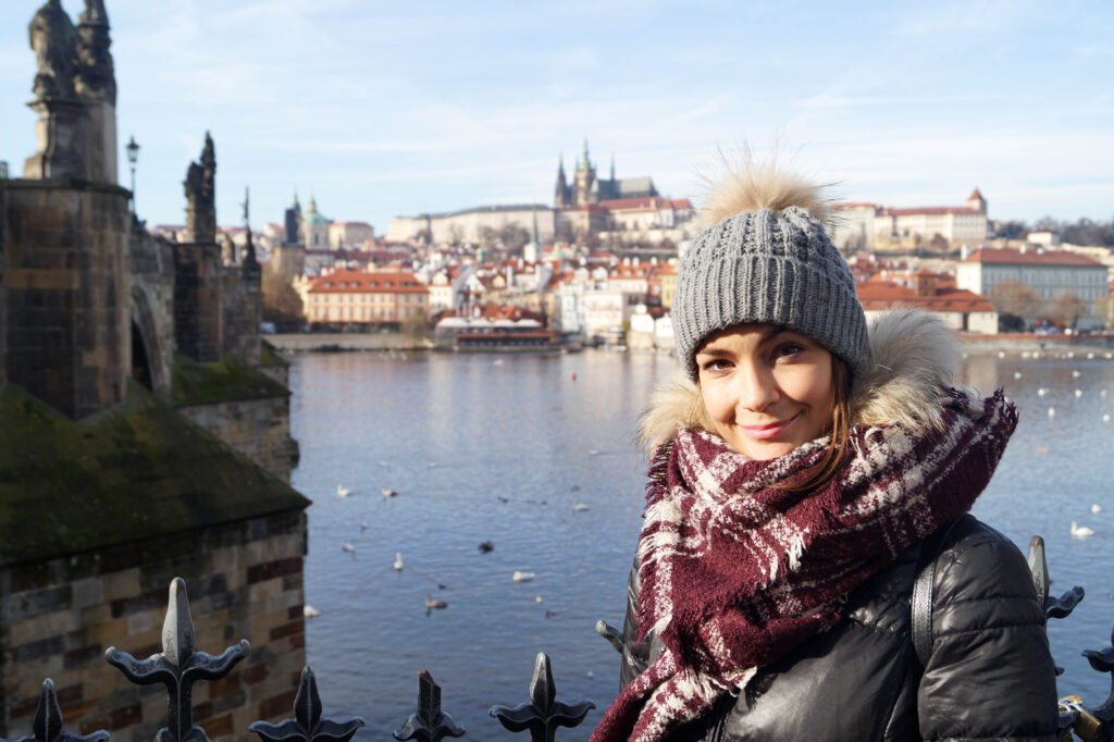 mujer con un sombrero gris, bufanda púrpura de pie frente al castillo de Praga, uno de los mejores puntos de fotos