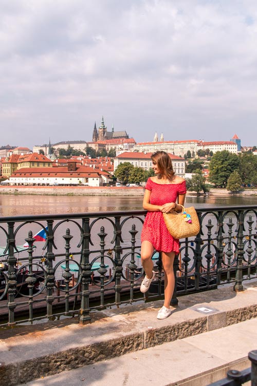 mujer con un vestido rojo de pie red a la lluvia negra con una vista sobre el río y el castillo de Praga