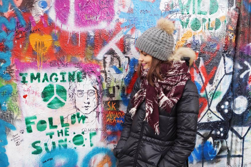 mujer con sombrero gris, chaqueta negra frente al colorido Muro joh lenon en Praga